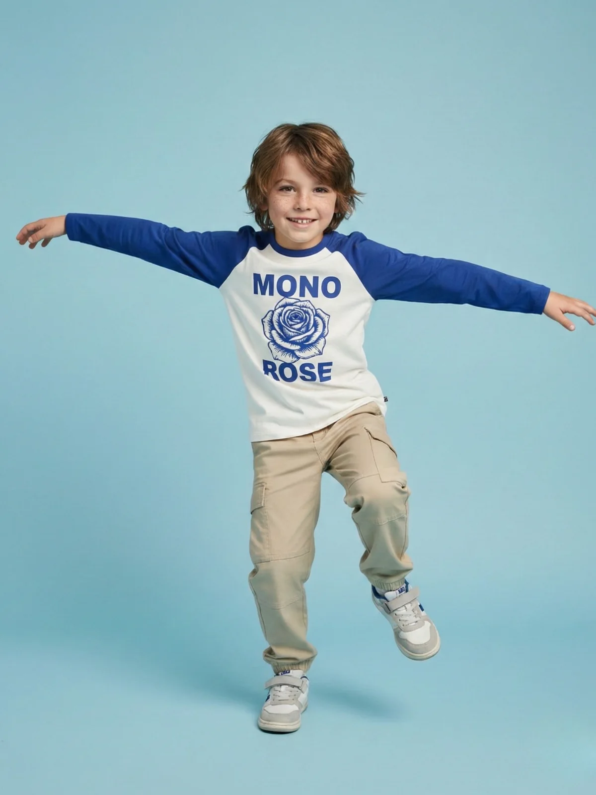 Chłopczyk stojący na niebieskim tle na jednej nodze delikatnie sie uśmiechajac, ma na sobie bezowe spodnie cargo, sportowe sneakersy oraz bluzke z roza