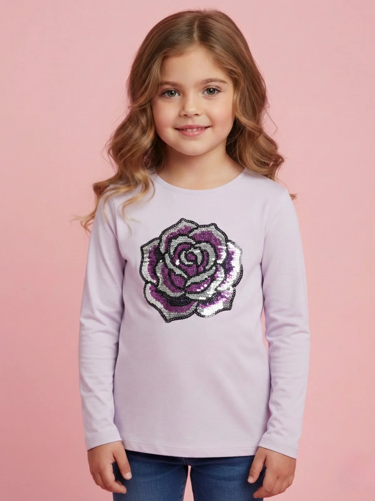 lekko uśmiechnięta dziewczynka w rozowej bluzeczce z ozdobną różą w kolorze srebrno różowym, stoi na różowym studyjnym tle