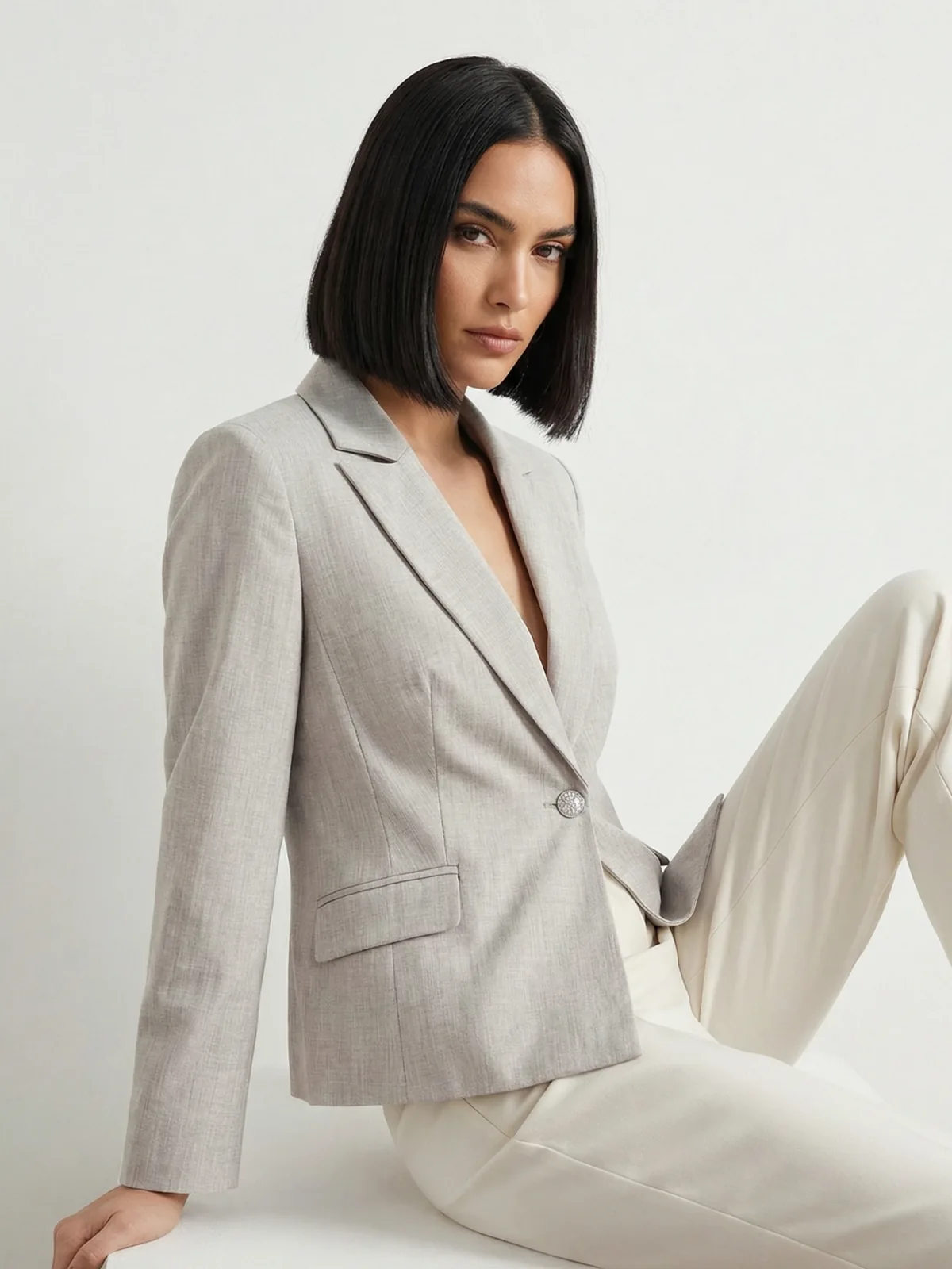Modelka o czarnych krótkich wlosach siedzi w jasnym studio ubrana w elegancką stylizacje z beżowego żakietu oraz białych spodni