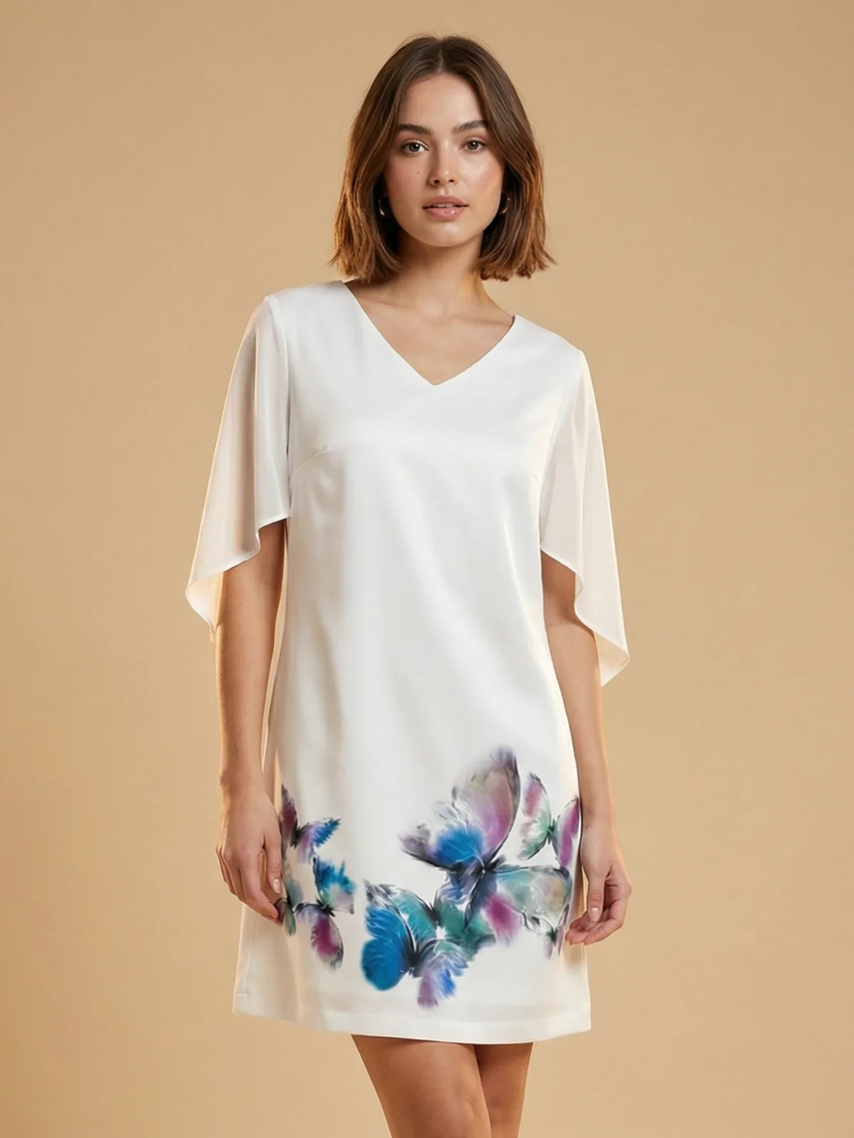 Młoda modelka prezentuję sukienkę w kolorze kremowym z charakterystycznym obrazem w niebiesko-fioletowe motyle na dole produktu, stoi na beżowym tle