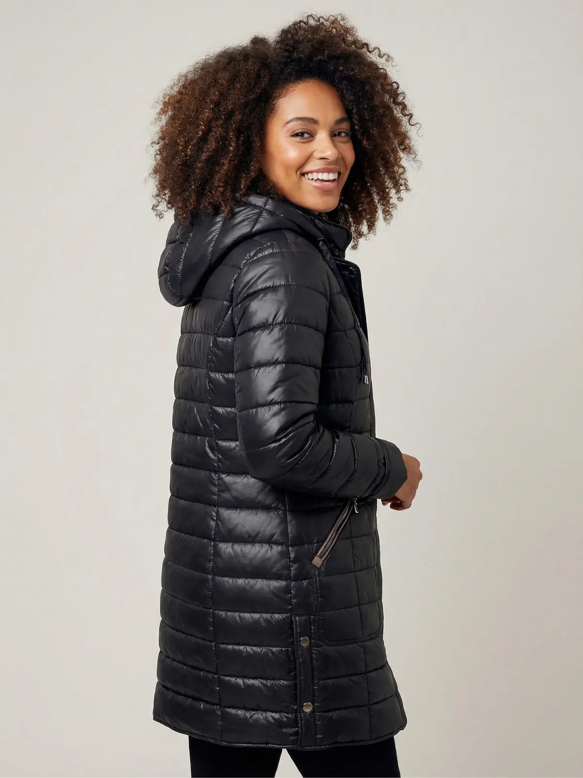 ujęcie z boku na modelke w kręconych włosach uśmiechająca się do obiektywu. ubrana w przejściową kurtkę w kolorze czarnym za kolano