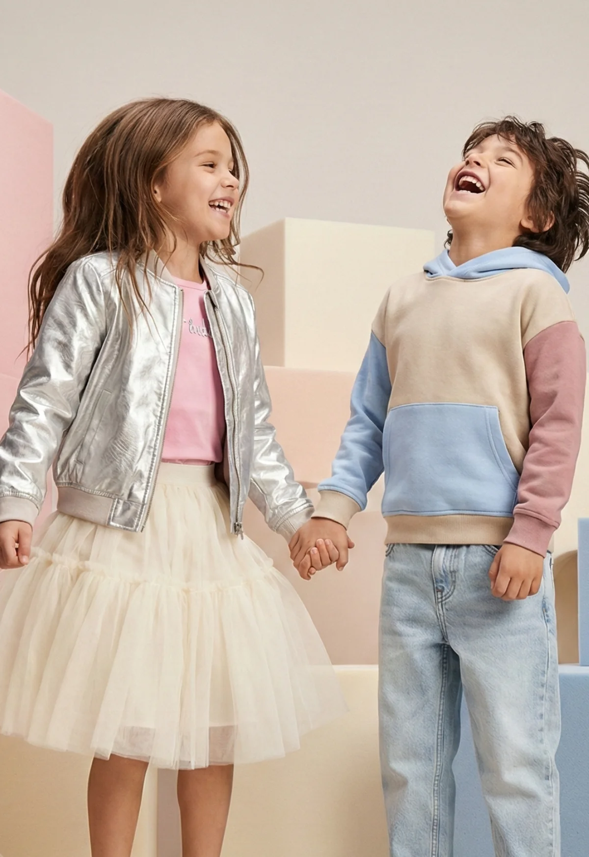 Dziewczynka i chłopec 7 lat prezetnują w studio nową kolekcje ubrań, uśmiechnięci skaczą do góry na tle z pastelowymi kwadratami