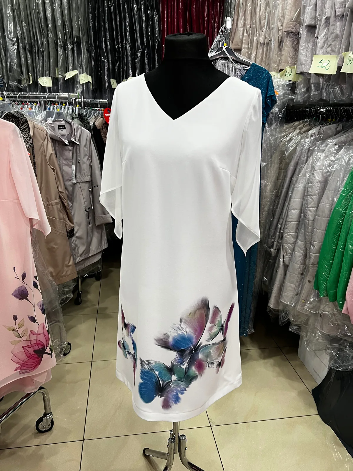 Biała sukienka elegancka w kolorze kremowym z nadrukiem w niebieskie motyle, zawieszona na manekinie w hurtowni polskiego producenta